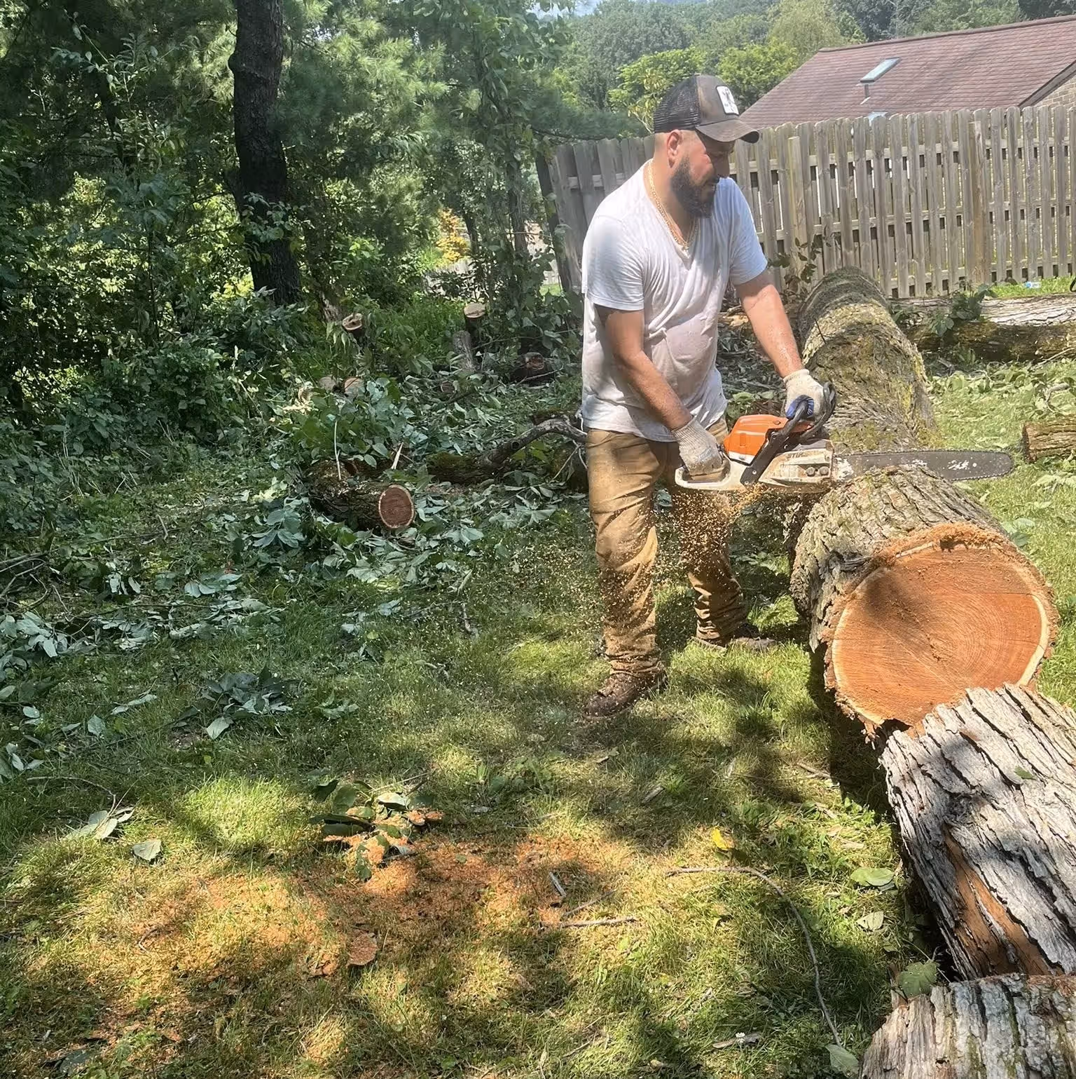 Man in white shirt and gloves cutting logs with a chainsaw in a leafy backyard.