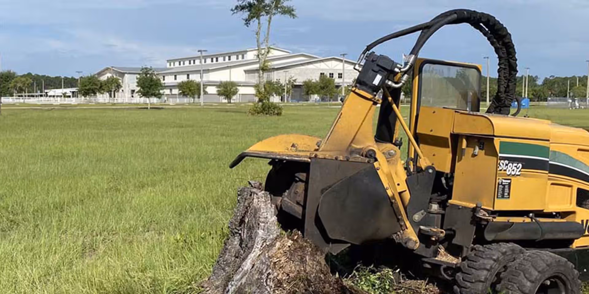 Yellow stump grinder machine attached to a large tree stump in a grassy field with buildings in background.